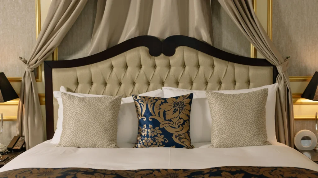 Illustrative stock image of a bed with a padded headboard, patterned cushions and draped curtains.