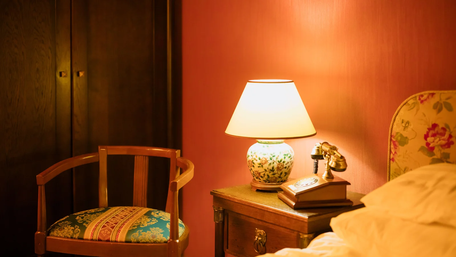 Illustrative stock image of a bedside lamp, telephone and wooden chair against a warm orange wall.
