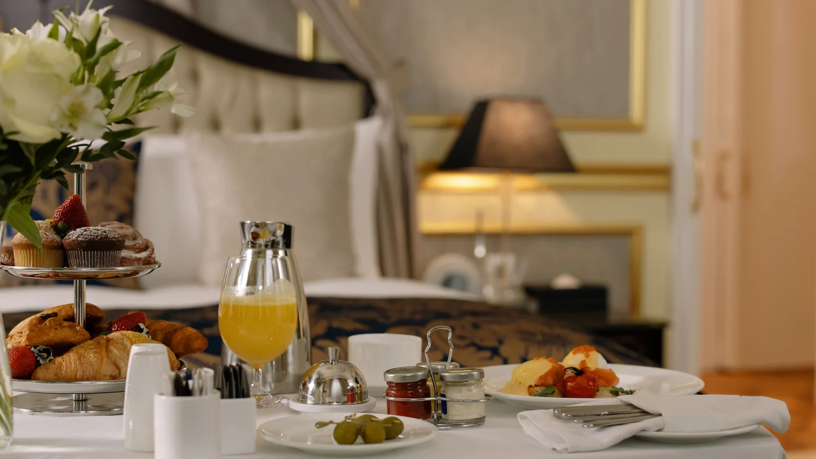 Breakfast table with pastries, orange juice, cups and flowers beside a bed.